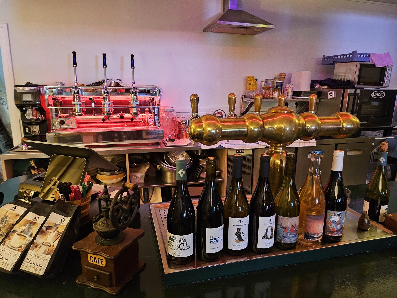 Le comptoir avec la machine à espresso, la tireuse en laiton, les bouteilles de vin et les sacs de café